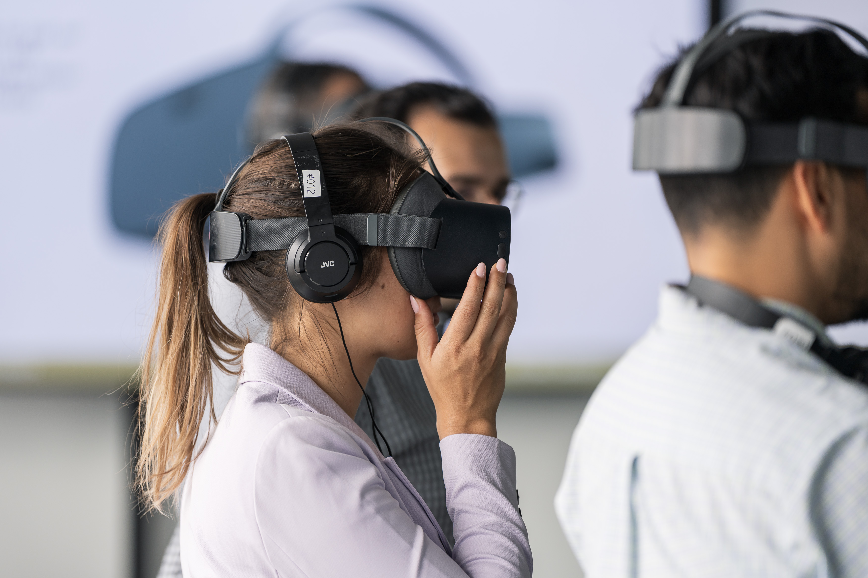 A young woman wearing a VR headset stands immersed in a virtual experience, one hand gently touching the device as she focuses on the simulation.